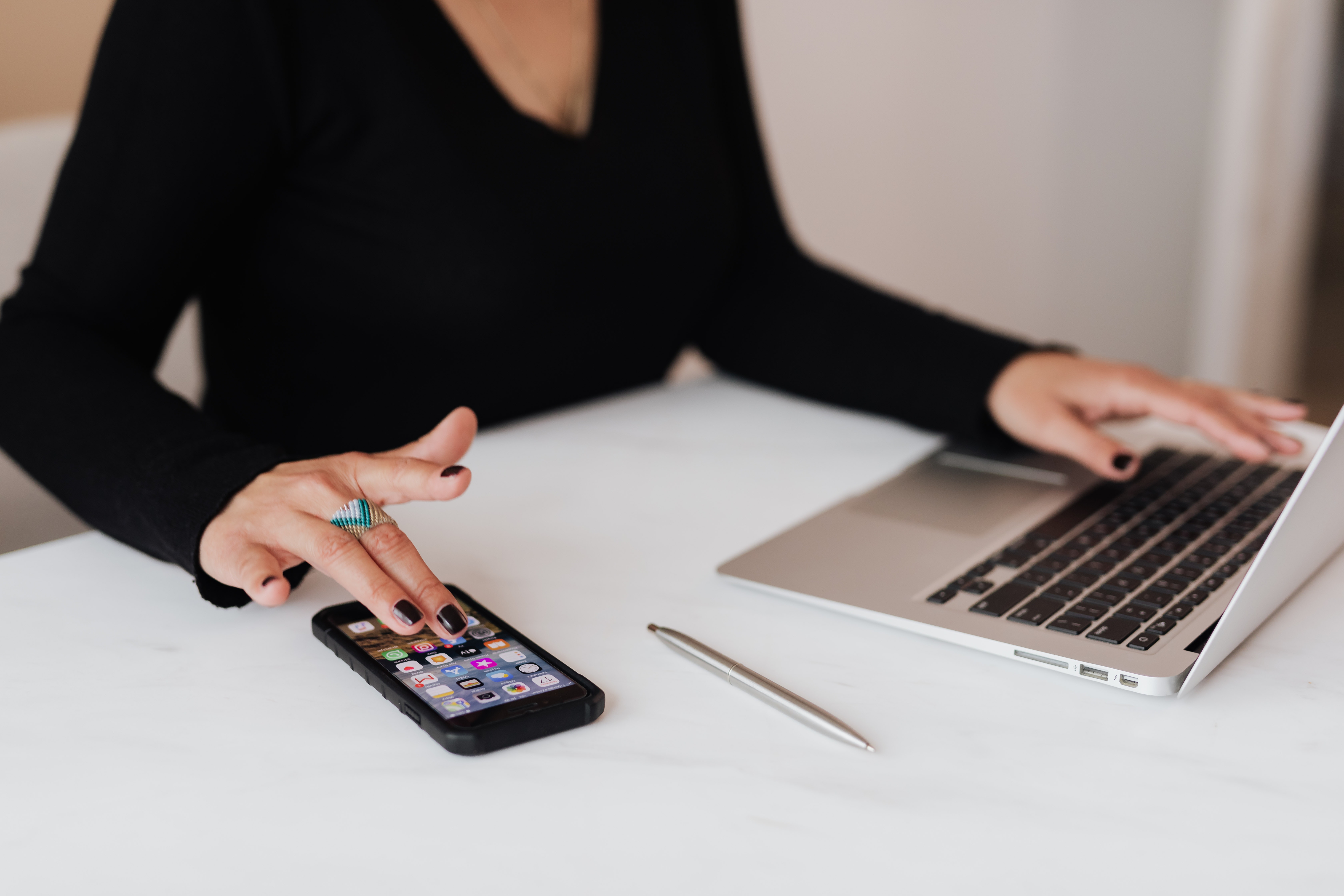 woman in long-sleeved v-neck top sitting with one hand on laptop keyboard and one hand on smartphone on a white surface with pen nearby