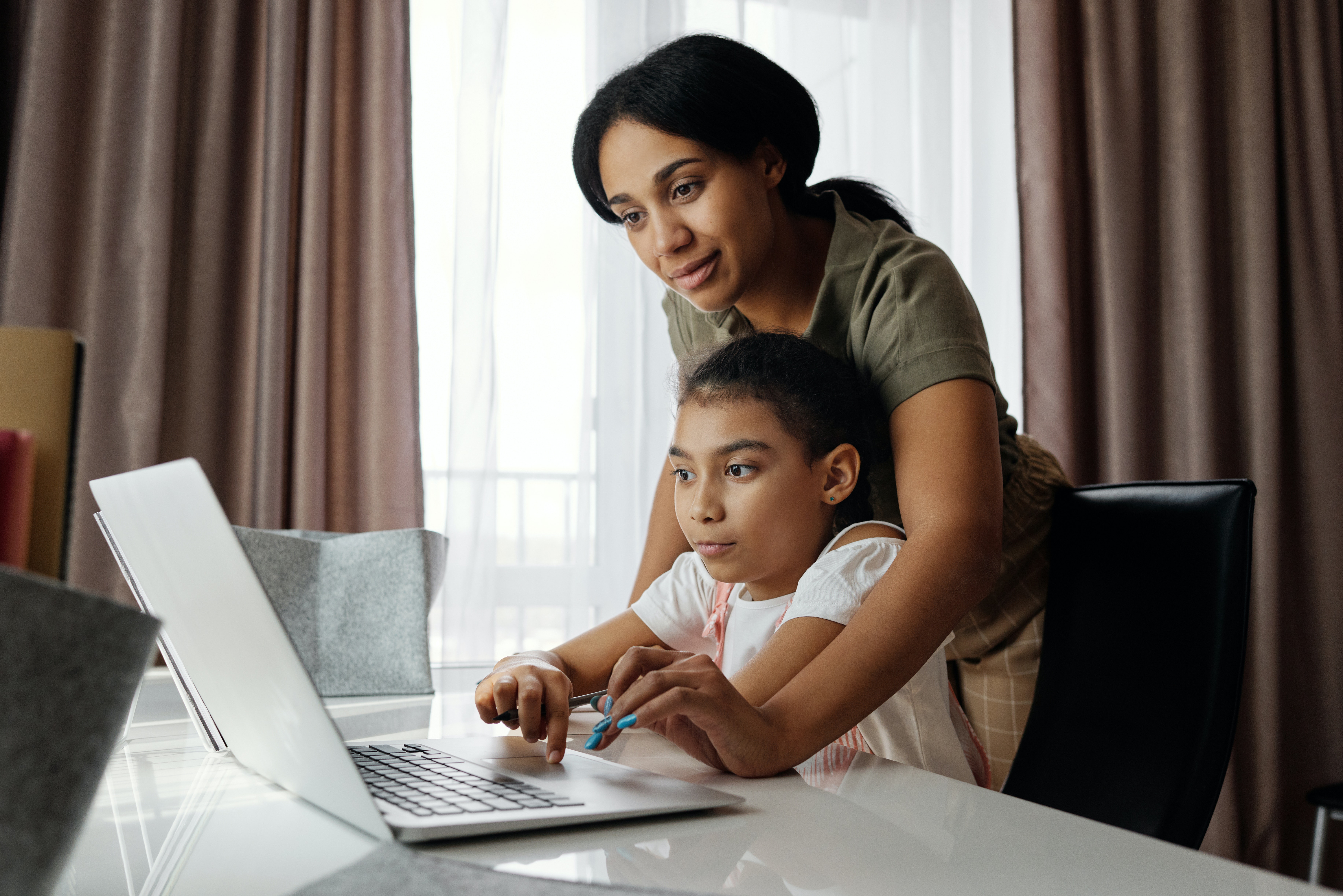 mother standing over daughter and helping her use laptop