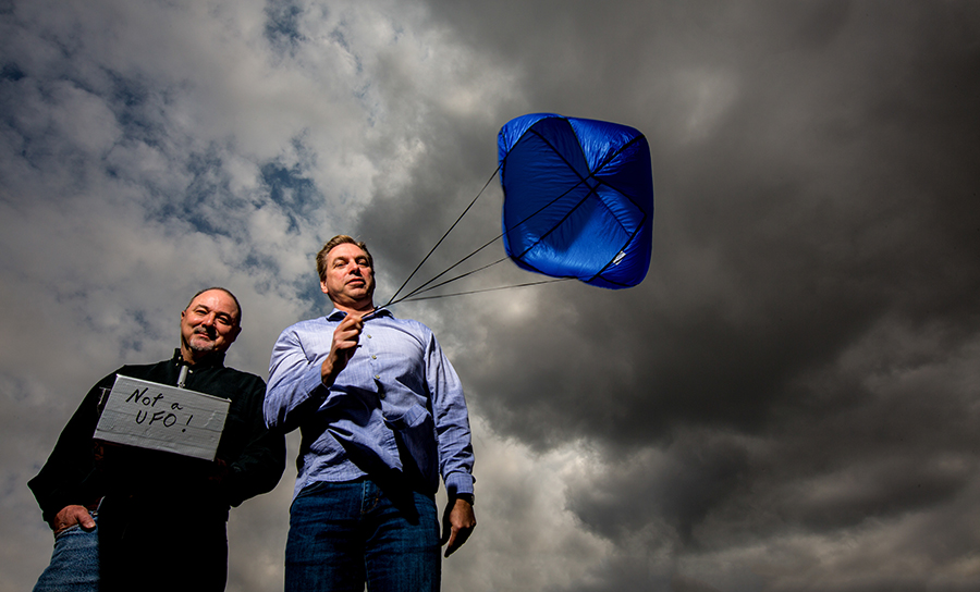 two men standing in front of a cloudy sky, one holding a board that reads "not a UFO" and the other holding a parachute